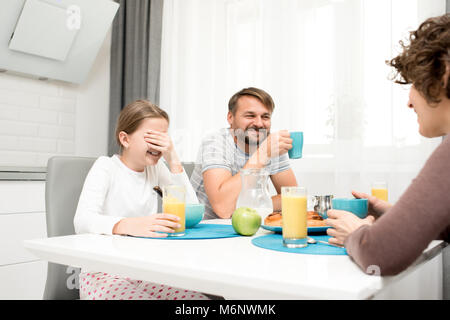 Portrait of carefree young family laughing happily while enjoying breakfast at home sitting at dinner table in cozy kitchen in morning, drinking orang Stock Photo