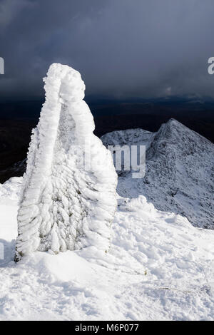 Ice covered marker stone at top of Watkin Path on Snowdon with Y Lliwedd beyond in winter snow in mountains of Snowdonia National Park Eryri. Wales UK Stock Photo