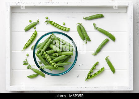 Pods of fresh green peas in glass bowl in white wooden tray, top view Stock Photo