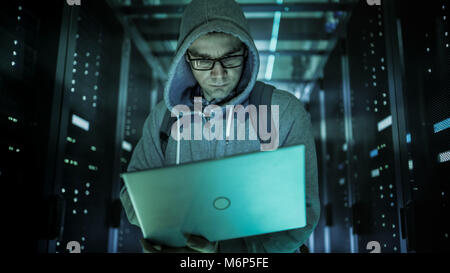Hacker in a Hoodie Standing in the Middle of Data Center full of Rack Servers and Hacking it with His Laptop. Stock Photo