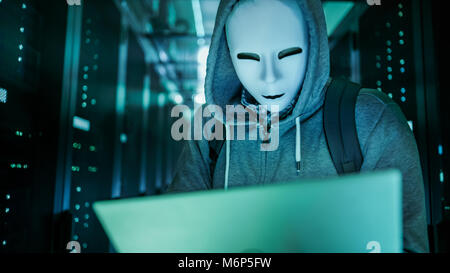 Mid Shot of a Masked Hacker in a Hoodie Standing in the Middle of Data Center full of Rack Servers and Hacking it with His Laptop. Stock Photo