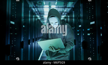 Mid Shot of a Masked Hacker in a Hoodie Standing in the Middle of Data Center full of Rack Servers and Hacking it with His Laptop. Stock Photo