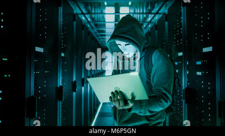 Mid Shot of a Masked Hacker in a Hoodie Standing in the Middle of Data Center full of Rack Servers and Hacking it with His Laptop. Stock Photo