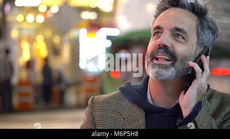 Happy successful businessman talking on mobile cell at night under bright lights of New York City in Times Square setting, smiling while holding phone Stock Photo