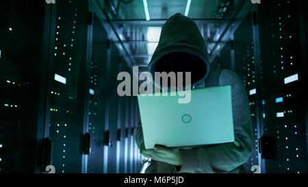 Close-up Shot of a Hacker with Hidden Face in a Hoodie Standing in the Middle of Data Center full of Rack Servers and Hacking it with His Laptop. Stock Photo