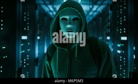 Masked Hacker in a Hoodie Standing in Corporate Data Center with Rows of Working Rack Servers. Stock Photo