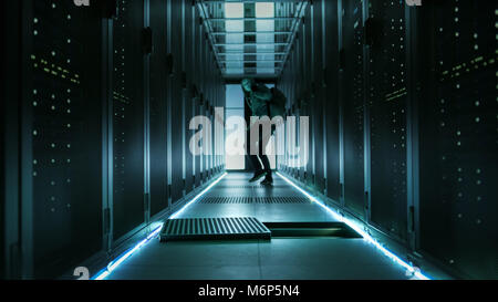 Shot of a Masked Hacker in a Hoodie Running Away and Watching Back with Laptop in a Corporate Data Center. Stock Photo