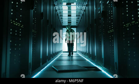 Hooded Hacker in a Mask Walks Through Working Data Center with Open Floor Hatch in the Middle of it. Stock Photo