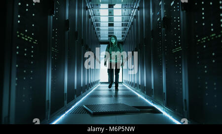 Hooded Hacker in a Mask Walks Through Working Data Center with Open Floor Hatch in the Middle of it. Stock Photo