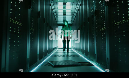 Hooded Hacker in a Mask Walks Through Working Data Center with Open Floor Hatch in the Middle of it. Stock Photo
