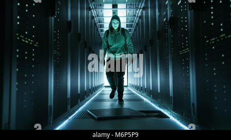 Hooded Hacker in a Mask Walks Through Working Data Center with Open Floor Hatch in the Middle of it. Stock Photo
