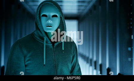 Masked Hacker in a Hoodie Watching at the Camera. He's in a Data Center. Stock Photo