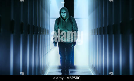 Masked Hacker Wearing Hoodies Walks in Through Data Center Corridor. Stock Photo