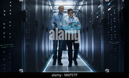 In Data Center Two IT Engineers Walking Through Rows of Server Racks. They Work on Tablet Computer and Laptop. Stock Photo