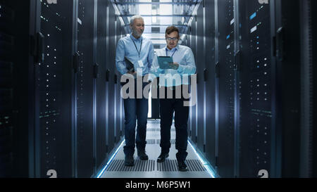In Data Center Two IT Engineers Walking Through Rows of Server Racks. They Work on Tablet Computer and Laptop. Stock Photo