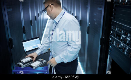 IT Engineer Taking Hard Drives off the Crash Cart and Installing Them into Rack Server. He's Working in Data Center. Also He Uses Laptop. Stock Photo