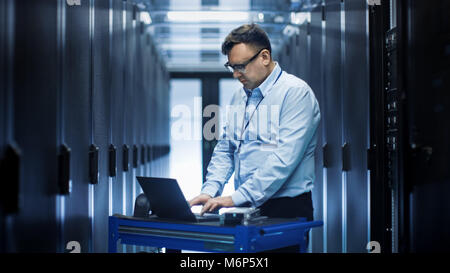 In Data Center IT Engineer Works on Crash Cart Laptop. We See Rows of Server Racks and One Cabinet is Open. Stock Photo