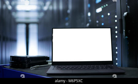 Laptop with White Screen on It Stands on The Table. In the Background Data Center With Rows of Server Racks and IT Engineer Working. Stock Photo