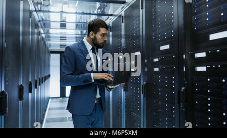 IT Engineer in Data Center Stands Before Server Rack Cabinet Working on His Laptop. Runs Diagnostics or Maintenance Job. Stock Photo