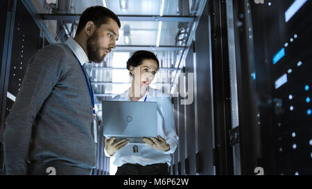Male Server Engineer is Looking at Server Rack Cabinet. His Female Colleague Works on a Laptop Beside Him. They're in Big Data Center. Stock Photo