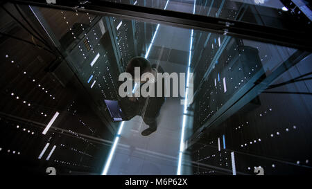 Top View Through the Glass of IT Engineer Working with Laptop in Data Center Full of  Active Rack Servers. Stock Photo