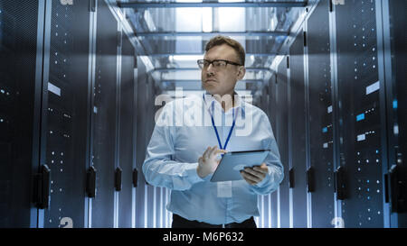 IT Technician Walks Through Rows of Server Racks in Data Center. Simultaneously He Works on a Tablet Computer. Stock Photo