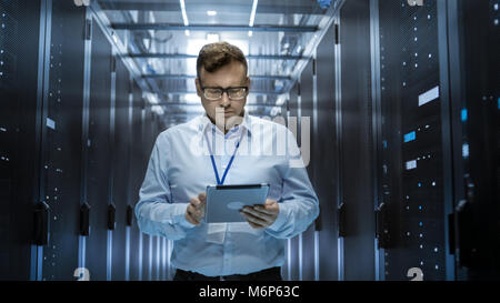 IT Technician Walks Through Rows of Server Racks in Data Center. Simultaneously He Works on a Tablet Computer. Stock Photo