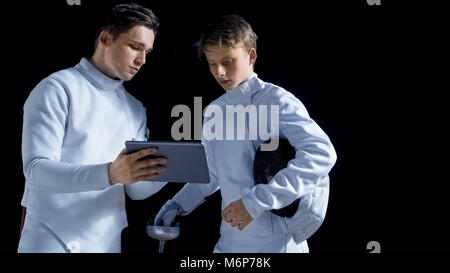 Two Young Fully Equipped Fencers Use Tablet Computer To Learn More About Strategy, Attack and Defense in Fencing. Shot Isolated on Black Background. Stock Photo