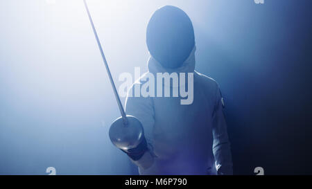 Fully Equipped Fencer Puts Lifts Foil Sword in Readiness for a Match. He Stands in the Spotlight while Darkness is Around Him. Shot Isolated on Black  Stock Photo