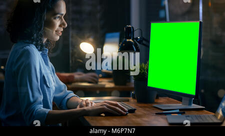 Beautiful Female Office Employee at Her Desk Works on a Mock-up Green Screen Personal Computer. Her Colleague Sits Beside. Creative Office Evening. Stock Photo