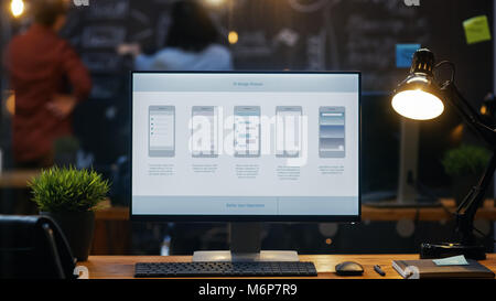 Personal Computer with Mobile Application Design Showing on the Monitor Stands on the Office Desk, In the Background People Work on a Blackboard Wall  Stock Photo