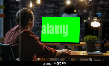 Office Employee at His Desktop Works on a Mock-up Green Screen Personal Computer. Over the Shoulder Footage. His Colleague works in the Background. Stock Photo