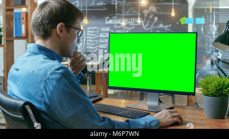 Young Man Works at His Desk on the Personal Computer with Mock-up Green Screen. In the Background His Colleague Works in the Creative Office. Stock Photo