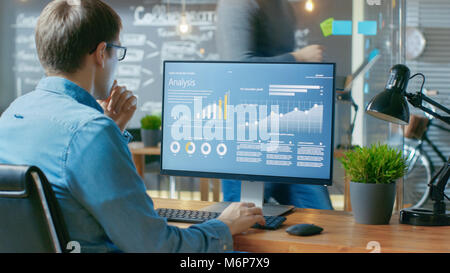 Analyst Works on a Personal Computer Showing Statistics, Graphs and Charts. In the Background His Coworker and Creative Office. Stock Photo
