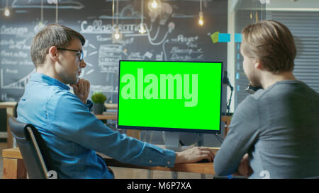 Two Office Employees Have Project Related Discussion, on the Desk Stands Personal Computer with Mock-up Green Screen. Stock Photo