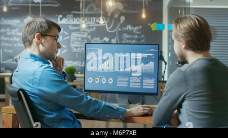 Two Office Employees Have Project Related Discussion, on the Desk Stands Personal Computer with Statistics, Graphs and Charts. Stock Photo