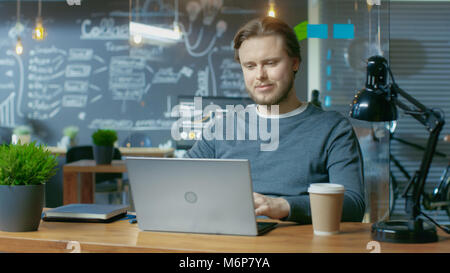 Handsome Young Office Employee Typing on a Laptop Computer,  He Charmingly Smiles. He's Working in the Creative Stylish Office. Stock Photo