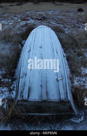 Upside down wooden boat on land covered with thin layer of snow. Spring year's grass in the background. Stock Photo