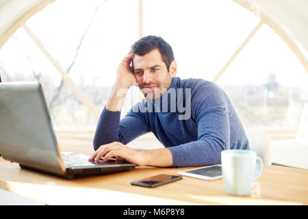 Portrait of young man with his forehead rested on his hand while sitting at desk and working on laptop. Home office. Stock Photo
