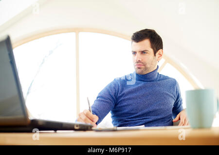 Portrait of casual young man wearing turtleneck sweater and looking thoughtul while working on laptop at home. Home office. Stock Photo