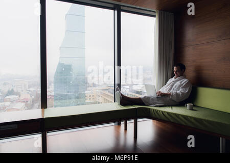 Businessperson working at home or in trip. Side view portrait of handsome young businessman sitting on bed and using laptop. Panoramic window with bea Stock Photo