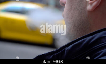 Young Millennial urban hipster blowing vapor electronic cigarette smoke outside on New York City street in day time. Yellow taxi cab passes in backgro Stock Photo