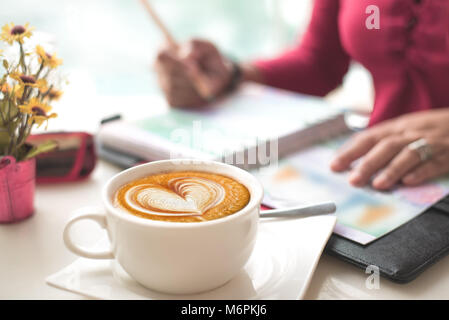 Coffee with Heart Shape Latte Art on Working Desk. Moody Shot, blur working woman checking figure in her chart in books. Concept passion work. Stock Photo