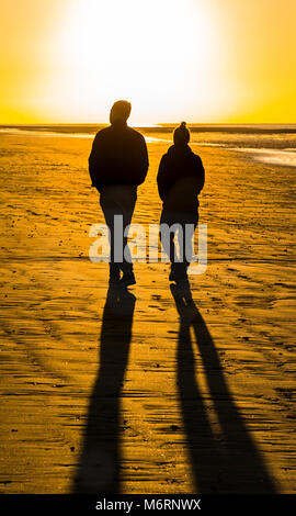 Couple of people walking on a sandy beach in the morning towards the rising sun, which casts long shadows. Stock Photo