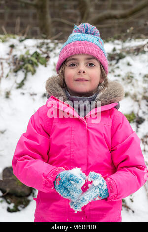 Pretty little girl playing in the snow during March winter holding a snowball and wearing a pink coat and blue pompom hat. Snowy weather in Cornwall Stock Photo