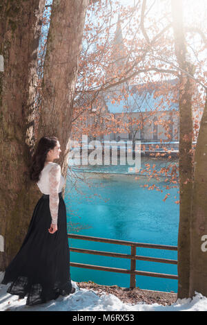 Young brunette woman, dressed in a black skirt, leaning against a tree, meditating on the shore of the Blautopf spring, in Blaubeuren, in Germany. Stock Photo