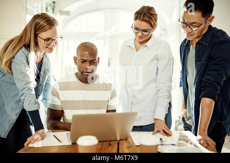 Diverse group of focused young design colleagues working together on a laptop at a table in a modern office Stock Photo