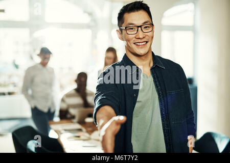 Smiling young Asian businessman extending a handshake while standing in an office with colleagues working in the background Stock Photo