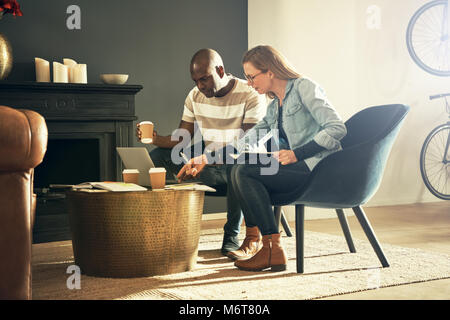 Two diverse young designers sitting together in a stylish modern office discussing a project and working online with a laptop Stock Photo