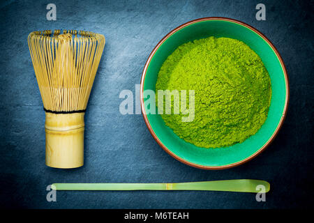 Matcha green tea powder in a bowl with bamboo whisk and spoon on dark slate background. Stock Photo